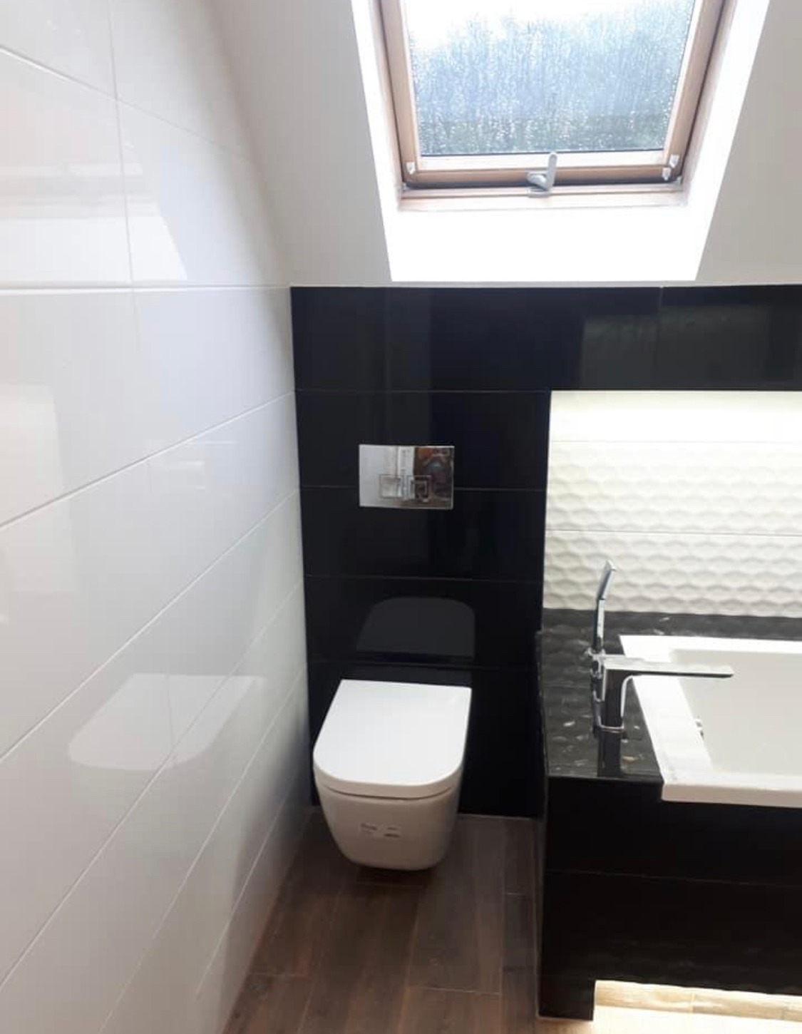 Nowoczesna łazienka na poddaszu z biało-czarnymi płytkami, podwieszaną toaletą i oknem dachowym.