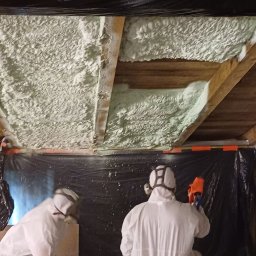 Usługi Remontowo - Budowlane Michał Wacław - Ocieplanie poddasza pianką PUR: Dwóch pracowników w kombinezonach ochronnych aplikuje pianę na drewniane elementy konstrukcyjne poddasza, zabezpieczone czarną folią.