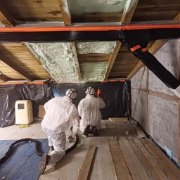 Usługi Remontowo - Budowlane Michał Wacław - Pracownicy w kombinezonach ochronnych aplikują jasną pianę izolacyjną na drewniany strop poddasza, zabezpieczonego czarną folią, widoczny agregat i przewody.