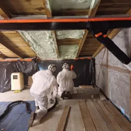 Pracownicy w kombinezonach ochronnych aplikują jasną pianę izolacyjną na drewniany strop poddasza, zabezpieczonego czarną folią, widoczny agregat i przewody.