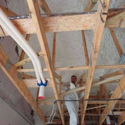 Usługi Remontowo - Budowlane Michał Wacław - Pracownik w kombinezonie zabezpieczającym aplikuje izolację na poddaszu, widoczne drewniane elementy konstrukcji dachu i instalacje. Praca na wysokościach.