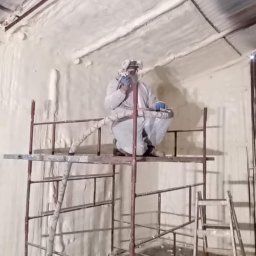 Usługi Remontowo - Budowlane Michał Wacław - Pracownik w kombinezonie na rusztowaniu, aplikuje pianę PUR na poddaszu w Kórniku. Widoczne ocieplone ściany i dach, oraz drabina. Ochrona dróg oddechowych.