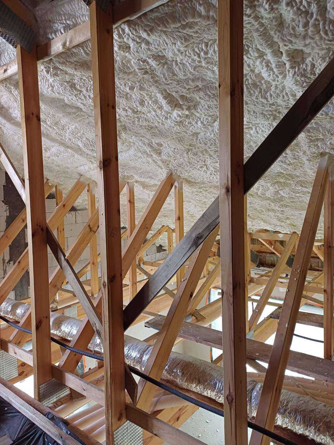 Ocieplenie poddasza pianką natryskową, widoczne drewniane elementy konstrukcyjne i instalacja wentylacyjna. Detal z bliska ukazuje strukturę pianki.