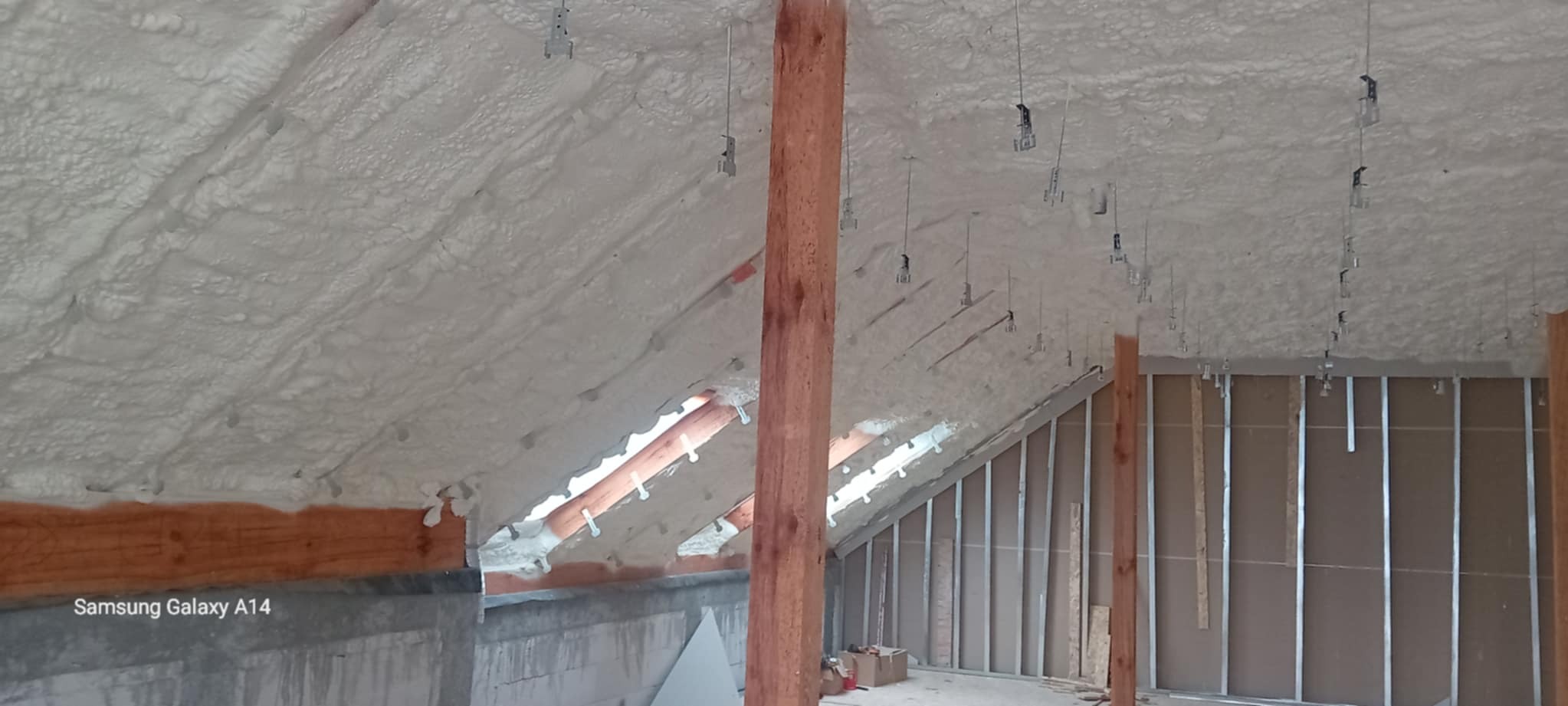 Poddasze po natrysku izolacji piankowej, widoczne drewniane elementy konstrukcyjne i metalowe uchwyty. Biała, nierówna powierzchnia piany pokrywa skośny dach.