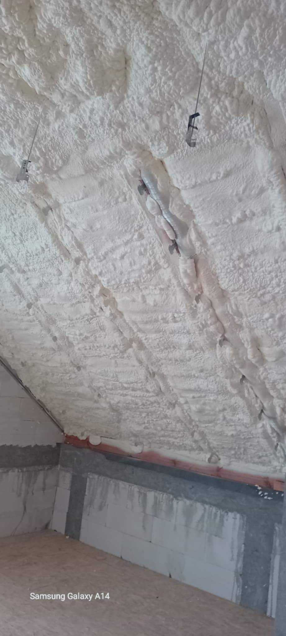 Ocieplenie poddasza pianką PUR: biała, nierówna powierzchnia piany pokrywa całą przestrzeń, widoczne elementy instalacji i fragment ściany.