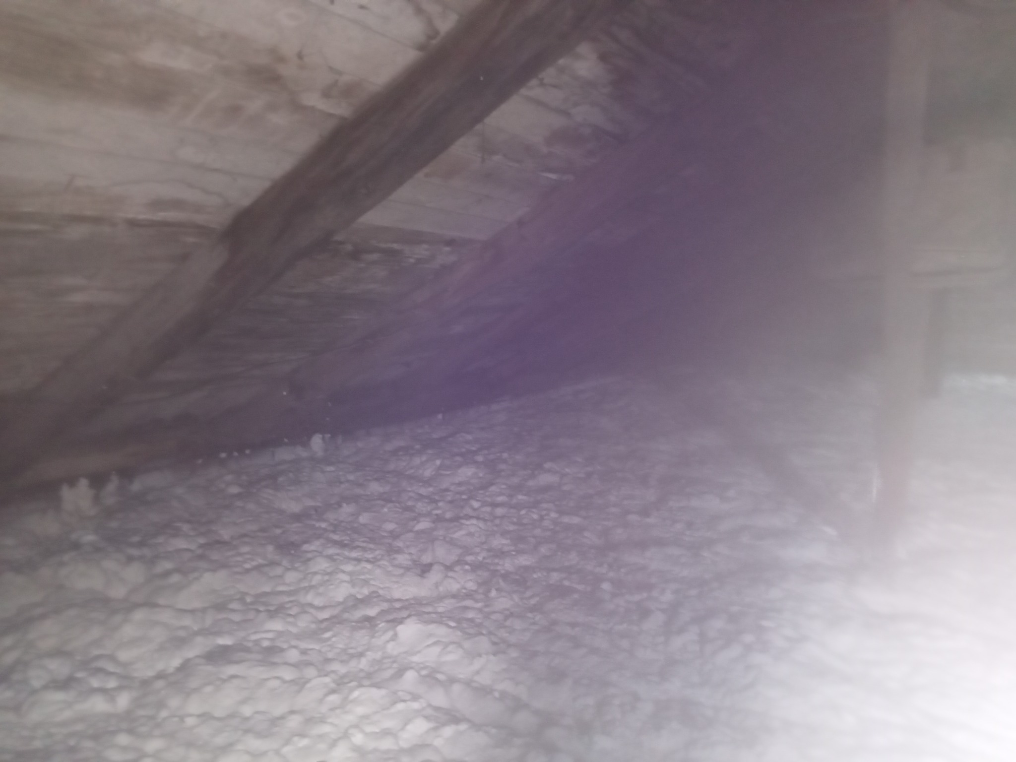 Izolacja poddasza pianą: biała warstwa pokrywa drewniane elementy konstrukcyjne. Nierównomierna powierzchnia piany, widoczne krople i struktura. Oświetlenie słabe, z lekką poświatą.