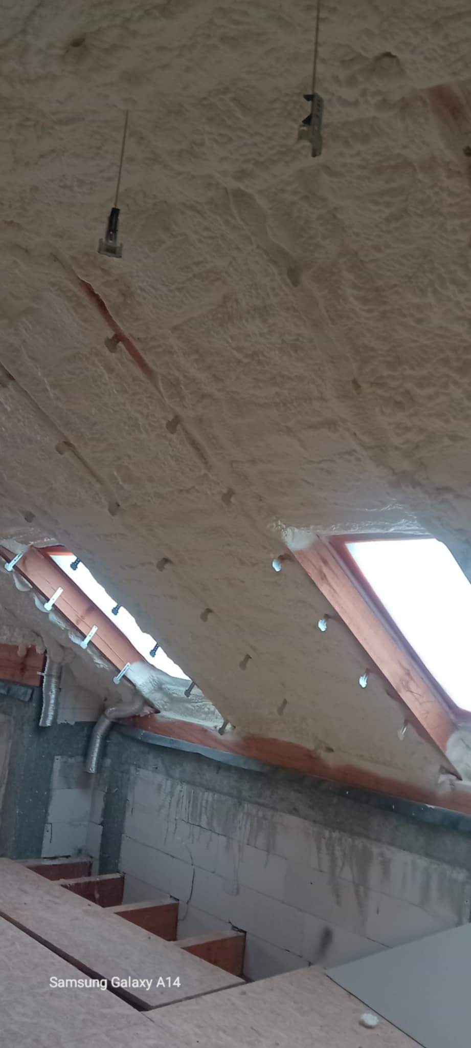 Ocieplenie poddasza pianką natryskową, widoczne krokwie i elementy montażowe. Biała, nierówna powierzchnia izolacji. Ujęcie z dołu, fragment okna dachowego.