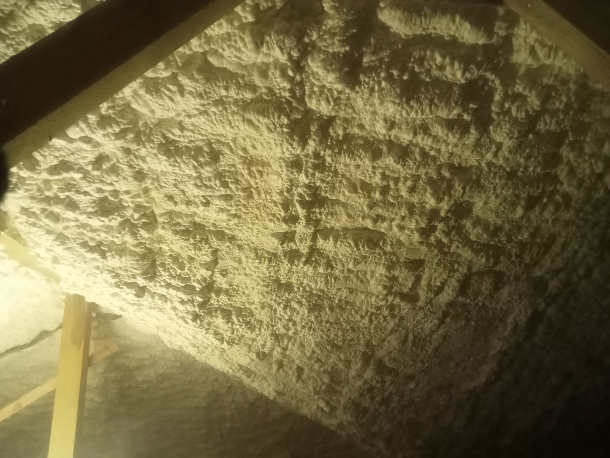 Ocieplenie poddasza pianką PUR, widoczne krokwie. Aplikacja pianki o nieregularnej strukturze, jasny odcień. Detal izolacji natryskowej w przestrzeni dachu.