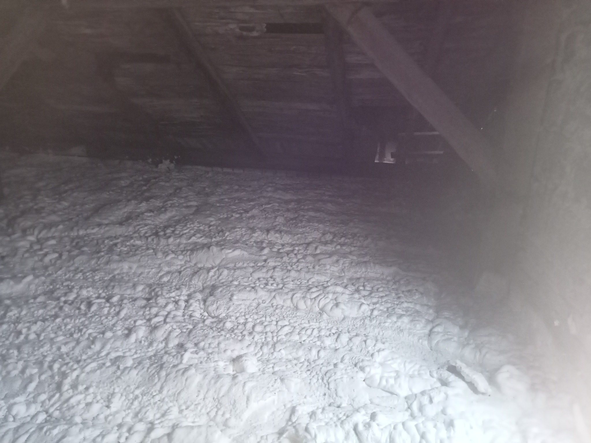 Szczegółowe ujęcie poddasza po aplikacji izolacji natryskowej. Widoczna struktura piany, drewniane elementy konstrukcyjne. Oświetlenie podkreśla nierówności powierzchni.