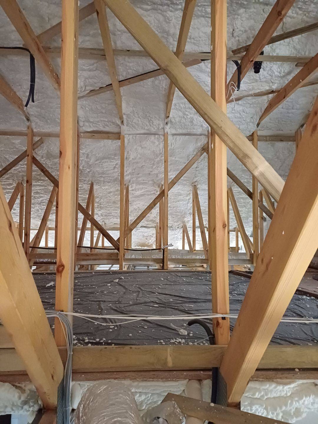 Ocieplenie poddasza pianką natryskową między drewnianymi krokwiami. Widoczna czarna folia izolacyjna na podłodze i biała pianka na skośnych powierzchniach dachu.