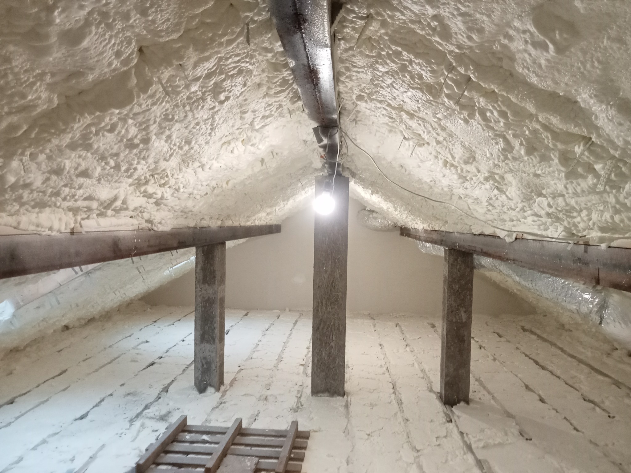 Poddasze po natrysku izolacji piankowej, widoczne drewniane elementy konstrukcyjne i instalacja wentylacyjna. Biała, nierówna powierzchnia piany pokrywa całą przestrzeń.