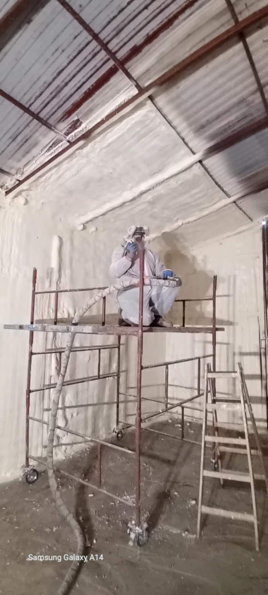 Pracownik w kombinezonie na rusztowaniu, aplikuje pianę PUR na poddaszu w Kórniku. Widoczne ocieplone ściany i dach, oraz drabina. Ochrona dróg oddechowych.