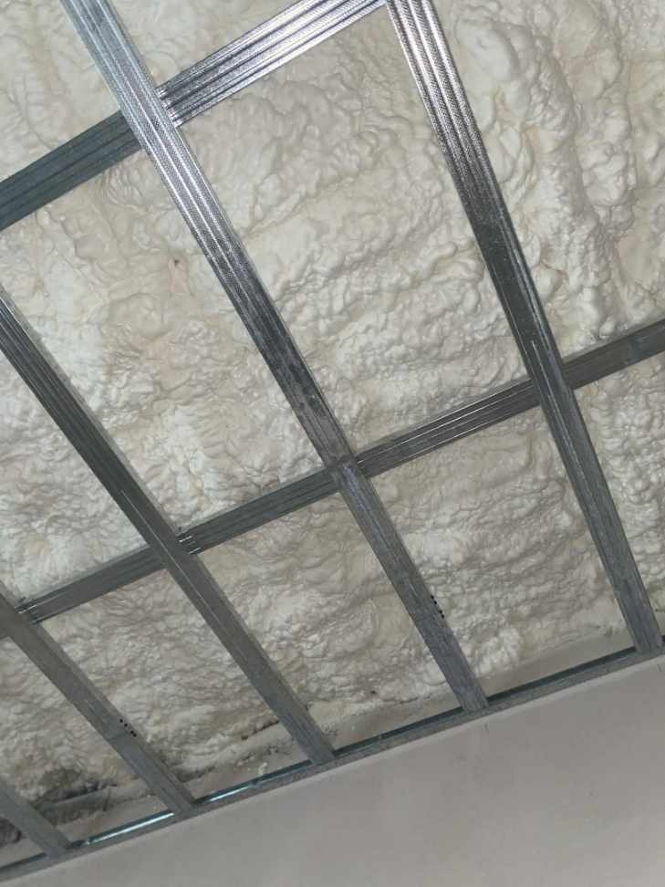 Ocieplenie poddasza pianką natryskową, widoczne metalowe profile konstrukcyjne. Biała, nierówna powierzchnia pianki wypełnia przestrzeń między profilami.