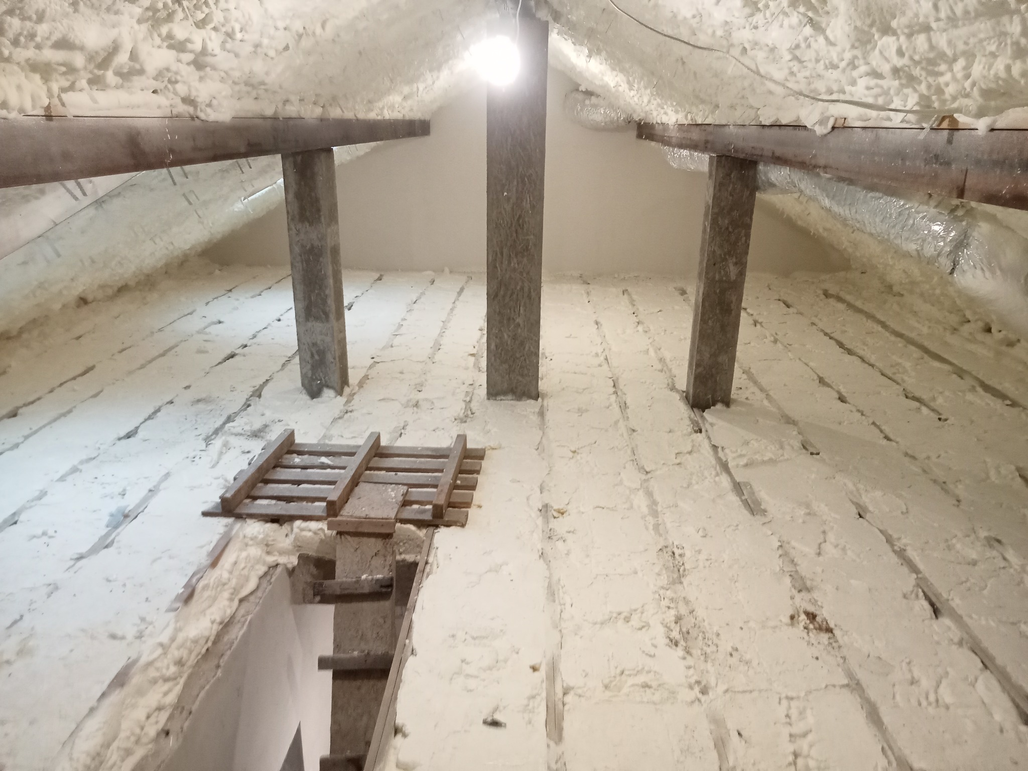 Poddasze po natrysku izolacji. Widoczne drewniane elementy konstrukcyjne i biała, nierówna powierzchnia pianki. Oświetlenie górne. Izolacja poddasza.