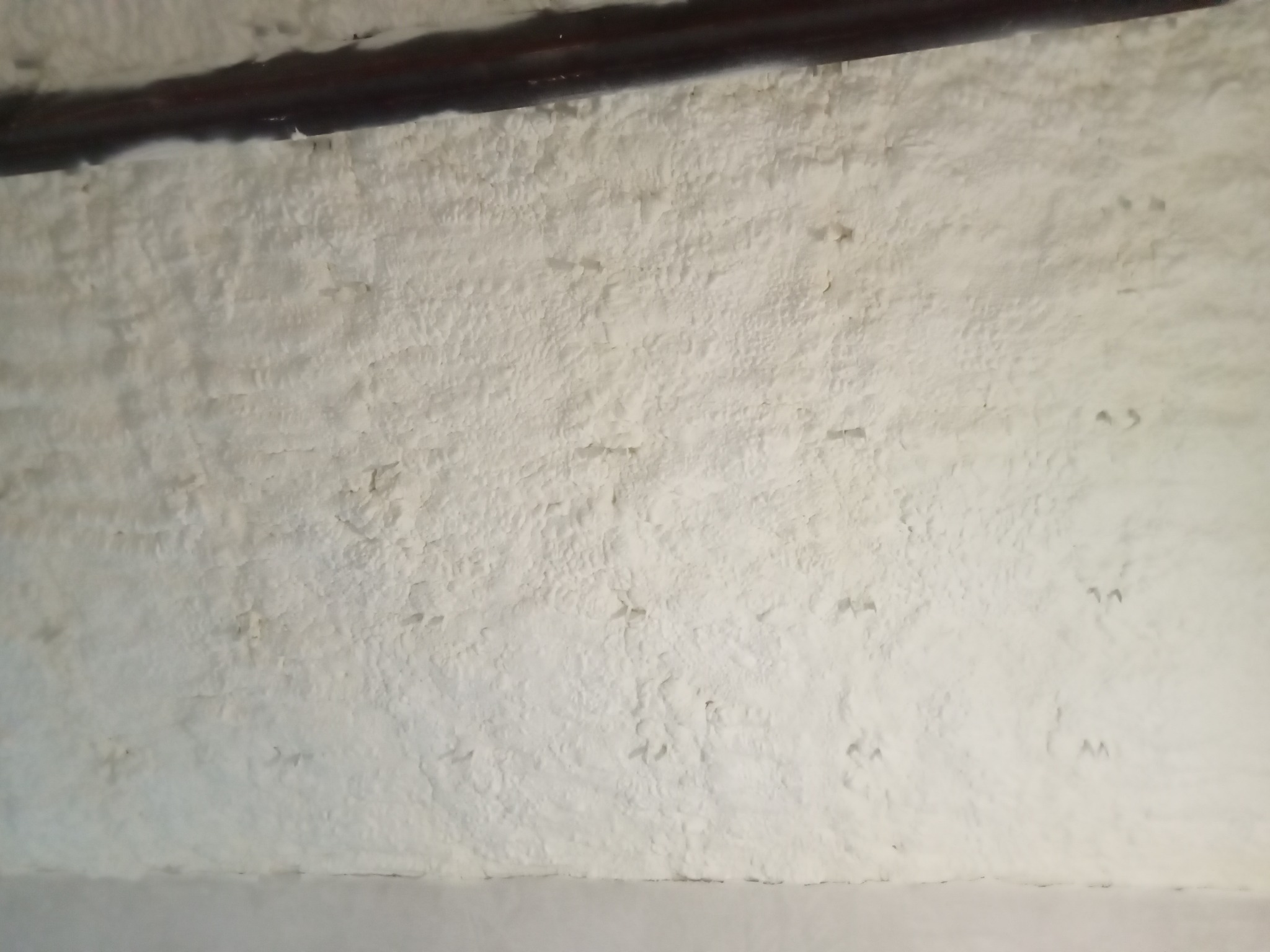 Szczegółowe ujęcie białej, nierównej powierzchni izolacji natryskowej na poddaszu, z widoczną drewnianą belką stropową w górnej części kadru.