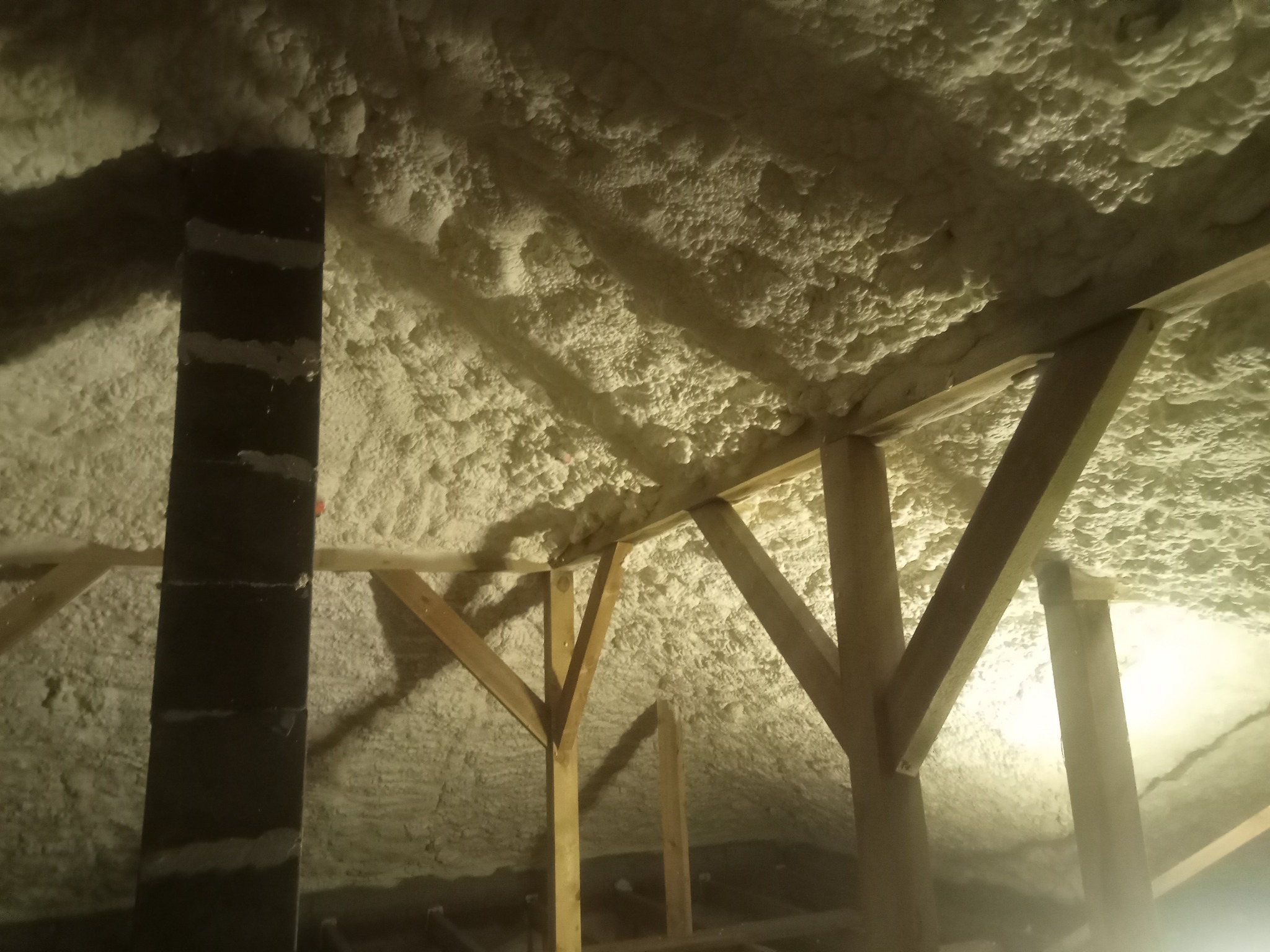 Poddasze po natrysku izolacji piankowej, widoczne drewniane elementy konstrukcyjne i komin. Aplikacja pianki pokrywa całą powierzchnię, tworząc jednolitą warstwę izolacyjną.