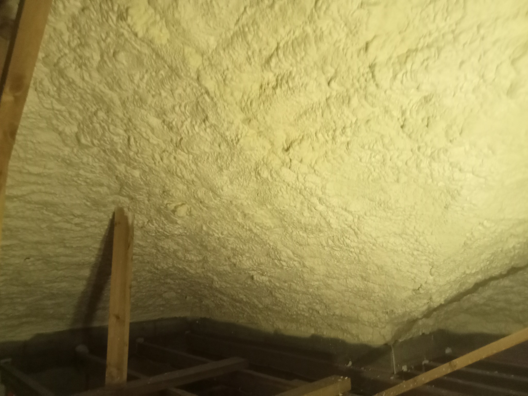 Ocieplenie poddasza pianką natryskową, widoczne drewniane elementy konstrukcji dachu. Żółta, nierówna powierzchnia pianki pokrywa całą przestrzeń.