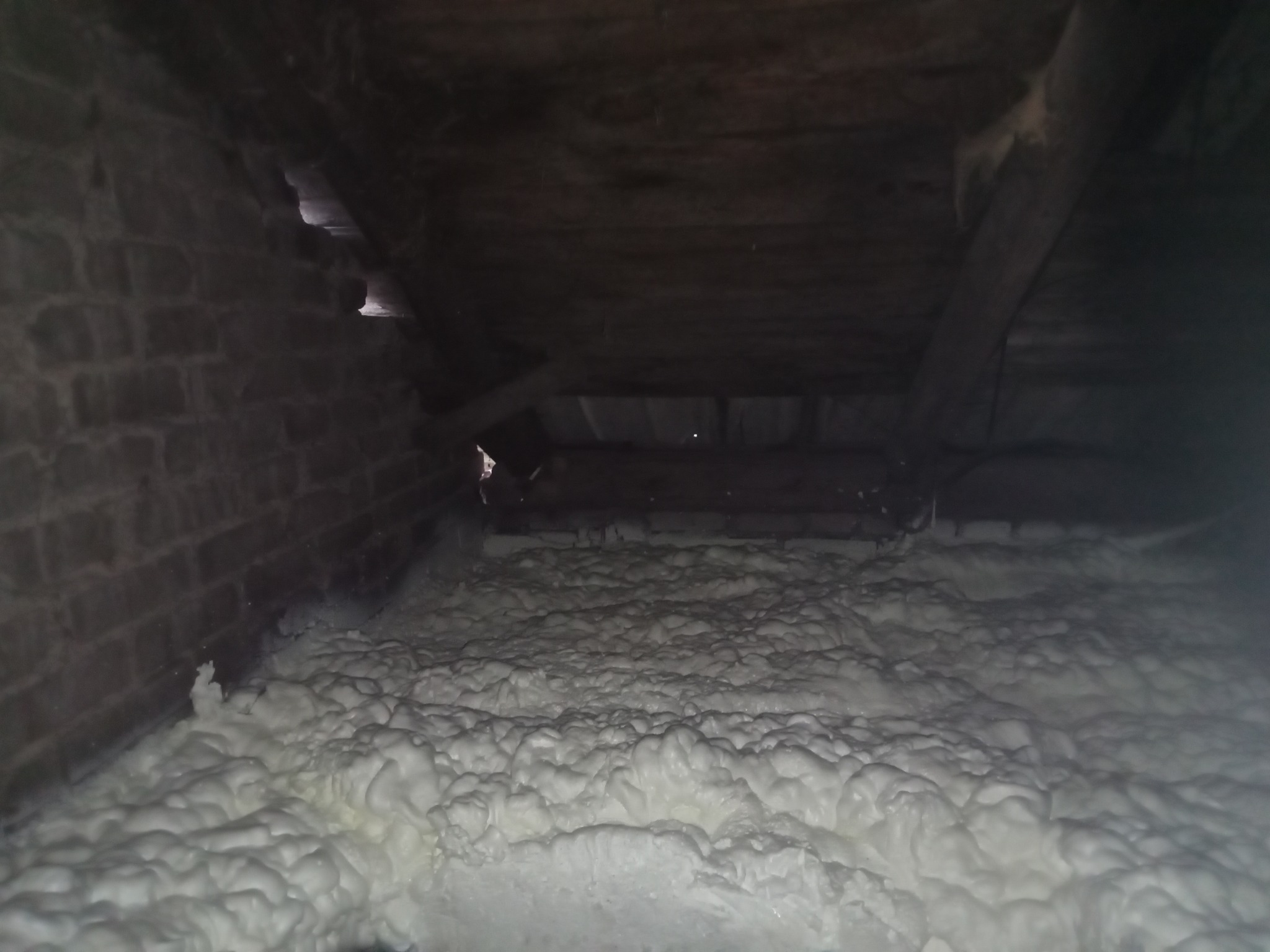 Poddasze z ceglanymi ścianami i drewnianą konstrukcją, wypełnione białą pianką PUR, ukazujące efekt ocieplenia. Widoczne nierówności i struktura pianki.