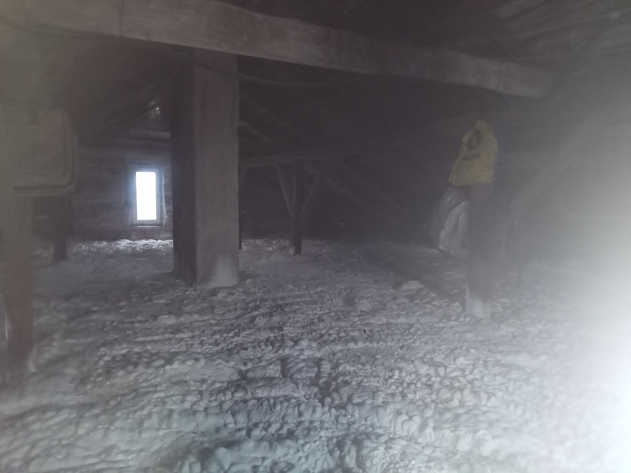 Ocieplone poddasze z widoczną warstwą izolacji natryskowej, drewnianą konstrukcją dachu i oknem w tle. Widoczne nierówności i struktura piany.