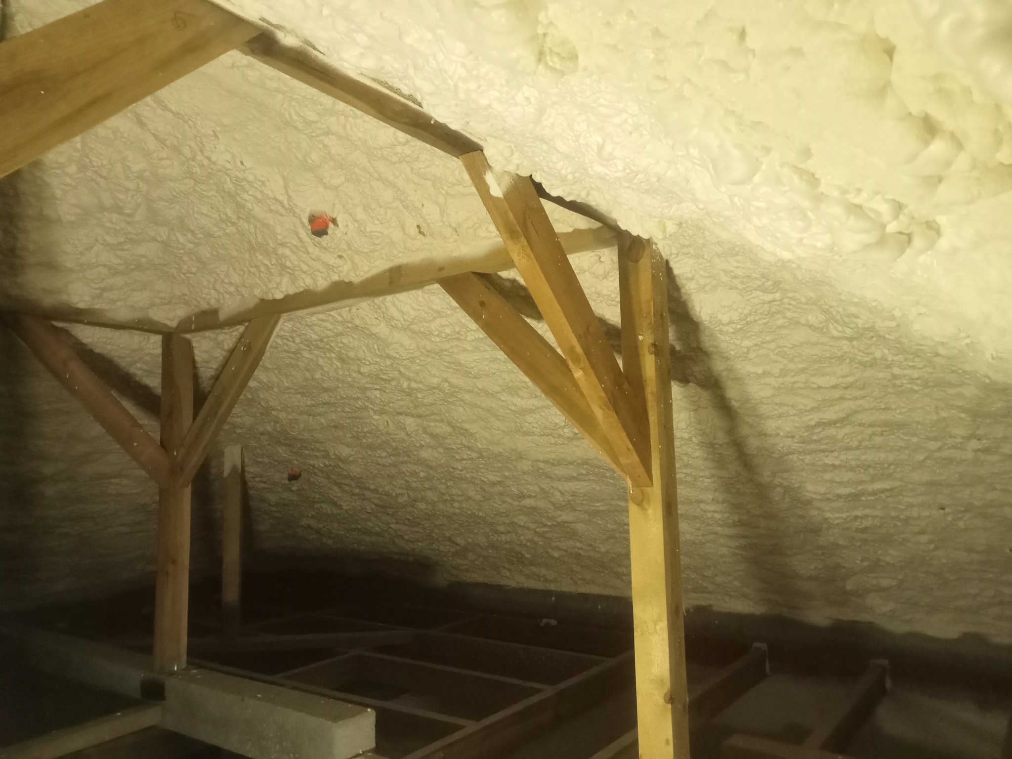 Poddasze z drewnianą konstrukcją pokryte grubą warstwą jasnej piany izolacyjnej, widoczne elementy więźby dachowej i podłogi. Izolacja natryskowa poddasza.