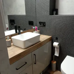 Nowoczesna łazienka z umywalką nablatową, drewnianym blatem i szarą mozaikową ścianą, widoczne akcesoria łazienkowe i toaleta.