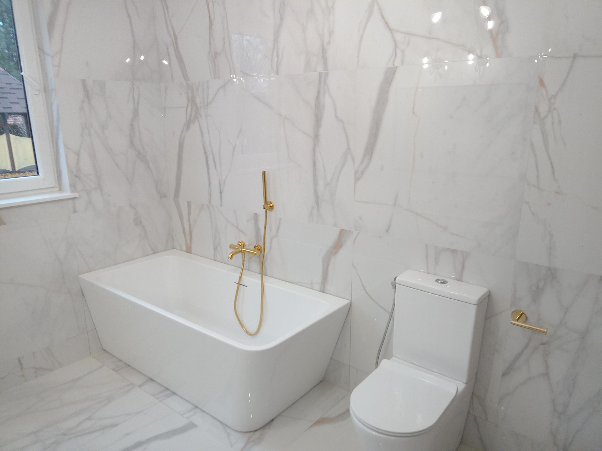 Nowoczesna łazienka w bieli i złocie: prostokątna wanna z baterią podtynkową, wisząca muszla klozetowa i płytki imitujące marmur Calacatta.