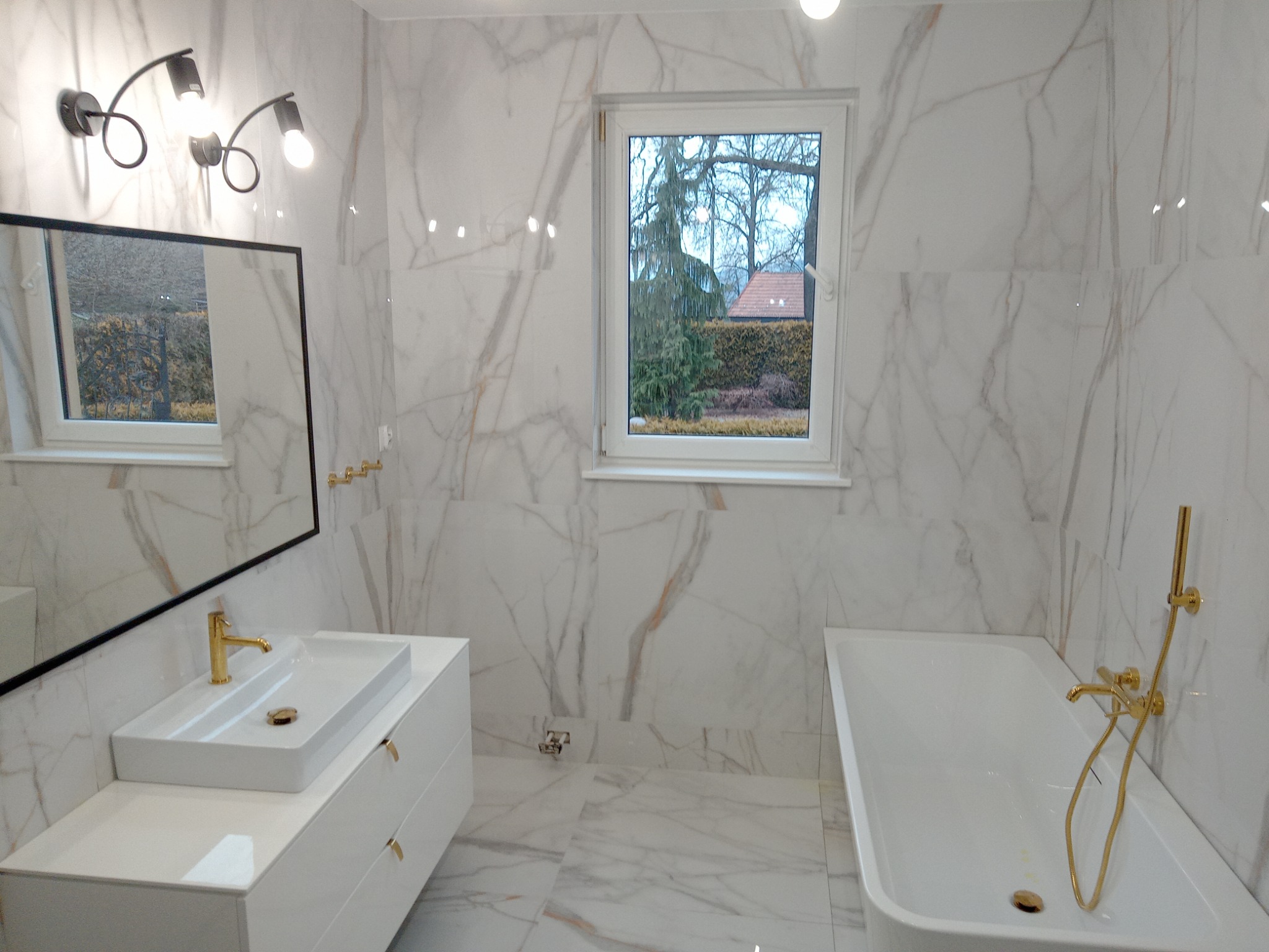 Nowoczesna łazienka z białymi marmurowymi płytkami, złotą armaturą, prostokątną umywalką na szafce i wanną, oświetlona lampą z trzema reflektorami.