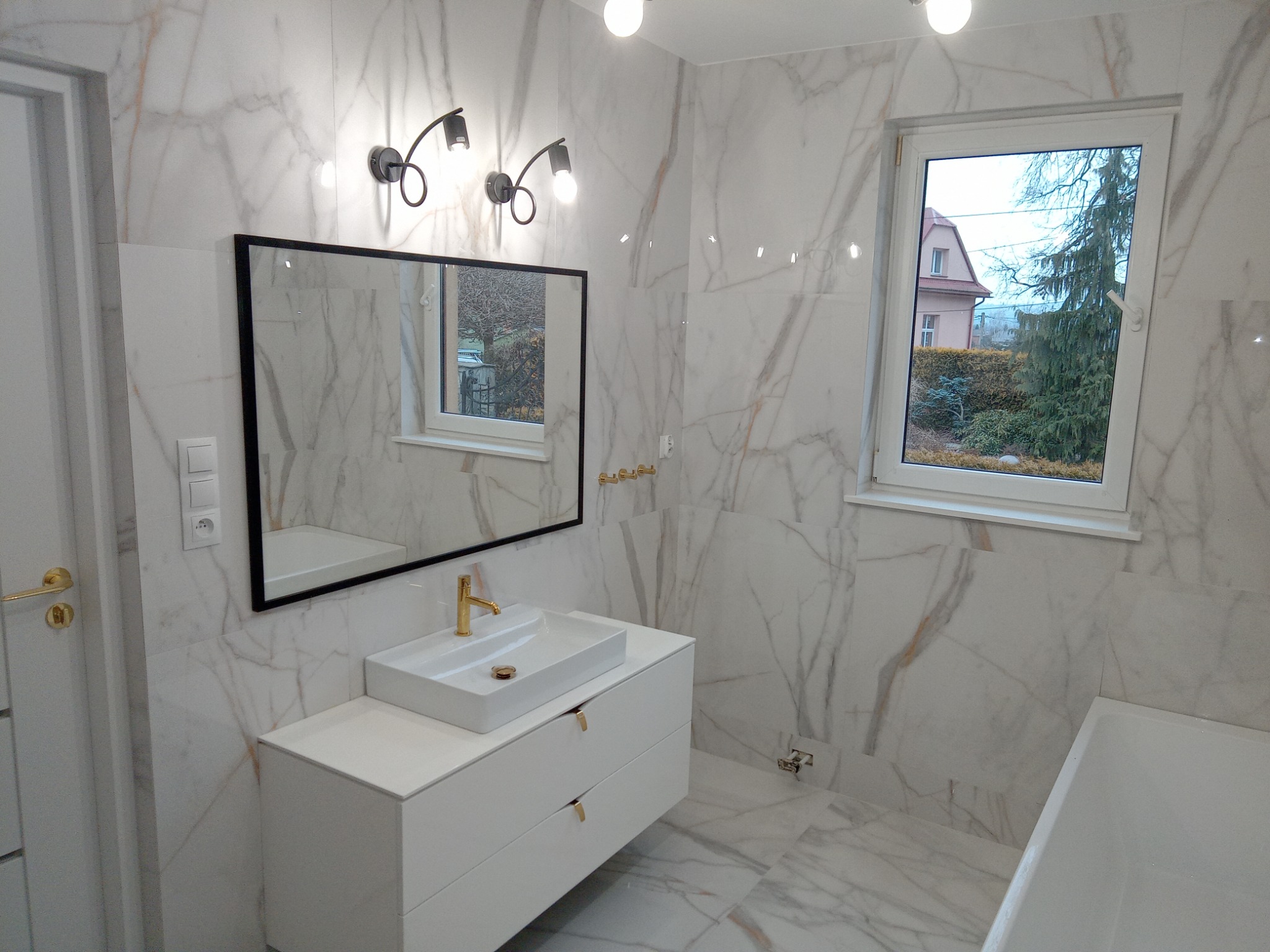 Nowoczesna łazienka z marmurowymi płytkami, białą szafką pod umywalką z złotą armaturą, czarnym lustrem i oknem z widokiem na ogród.