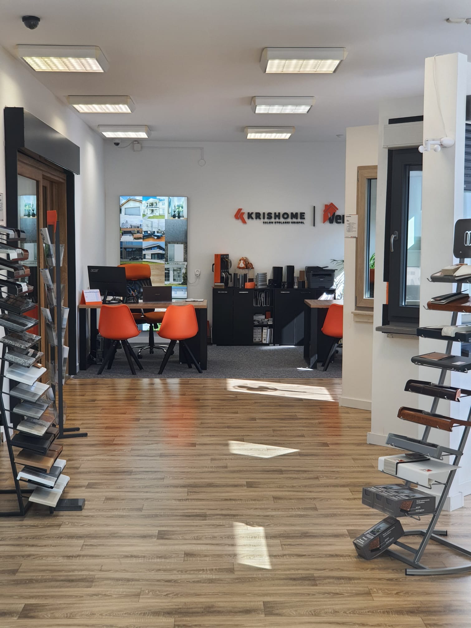 Wnętrze salonu stolarki okiennej i drzwiowej z ekspozycją próbek kolorystycznych na stojakach, widoczne biurka z komputerami i pomarańczowymi krzesłami oraz logo firmy KrisHome na ścianie.