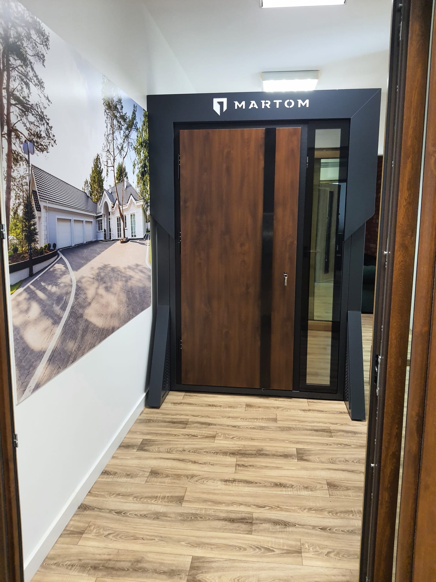 Wystawa drzwi zewnętrznych z drewnopodobnym wykończeniem i czarną ramą, obok ściana z fototapetą przedstawiającą dom jednorodzinny, widoczne logo producenta Martom.
