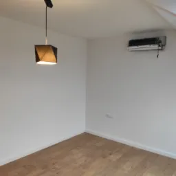 Puste, białe wnętrze poddasza z drewnianą podłogą, nowoczesną lampą wiszącą z czarnym abażurem i zamontowanym klimatyzatorem.