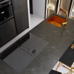 Nowoczesna kuchnia z ciemnymi szafkami, blatem z kamienia, złotą baterią przy miedzianym zlewie, płytą indukcyjną zintegrowaną z okapem i złotymi hokerami.