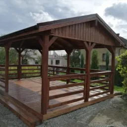 Zadaszona drewniana konstrukcja z barierkami, prawdopodobnie altana, z brązowym dachem i podłogą, otoczona zielenią i budynkami mieszkalnymi.