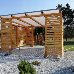 Nowoczesna konstrukcja z drewna z przezroczystym zadaszeniem, ustawiona na żwirowym podłożu, z widocznymi dwoma drewnianymi drabinami w tle.