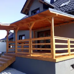 Drewniany taras z zadaszeniem przy szarym domu, z drewnianymi schodami i osobą pochyloną na schodach po lewej stronie.