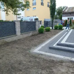 Nowoczesny, grafitowy płot panelowy z betonowymi słupkami, świeżo ułożona kostka brukowa z białymi obramowaniami, przygotowany teren pod trawnik, w tle budynki mieszkalne i osoby.