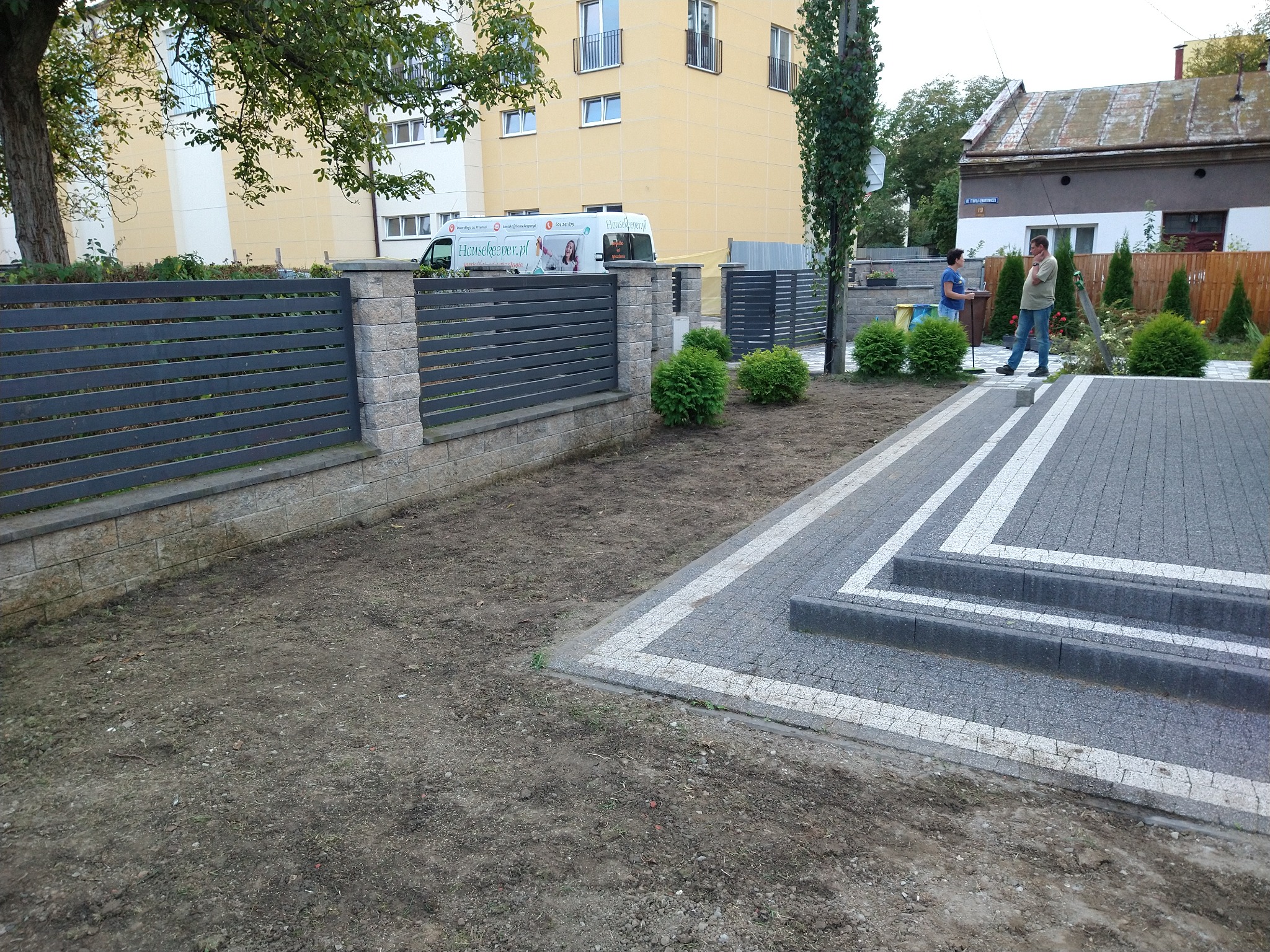 Nowoczesny, grafitowy płot panelowy z betonowymi słupkami, świeżo ułożona kostka brukowa z białymi obramowaniami, przygotowany teren pod trawnik, w tle budynki mieszkalne i osoby.