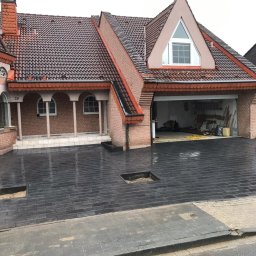 DANUTA PAWŁOWSKA ALFA-BRUK-2 - Ułożona, mokra, grafitowa kostka brukowa na podjeździe przed domem z odsłoniętymi fragmentami podłoża w dwóch miejscach, widoczny pracownik z maszyną wibracyjną, brama garażowa otwarta.