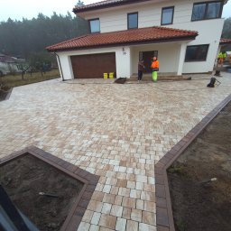 DANUTA PAWŁOWSKA ALFA-BRUK-2 - Nowo ułożona kostka brukowa w odcieniach beżu i brązu przed nowoczesnym domem z brązową bramą garażową, dwóch pracowników w odblaskowych ubraniach podczas czyszczenia kostki, widoczne elementy...