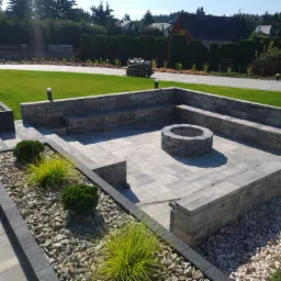 Ogród z paleniskiem otoczonym kamienną konstrukcją z siedziskami, wyłożony kostką brukową w odcieniach szarości, z fragmentami rabat wysypanych kamieniami i obsadzonych roślinami ozdobnymi, na tle...