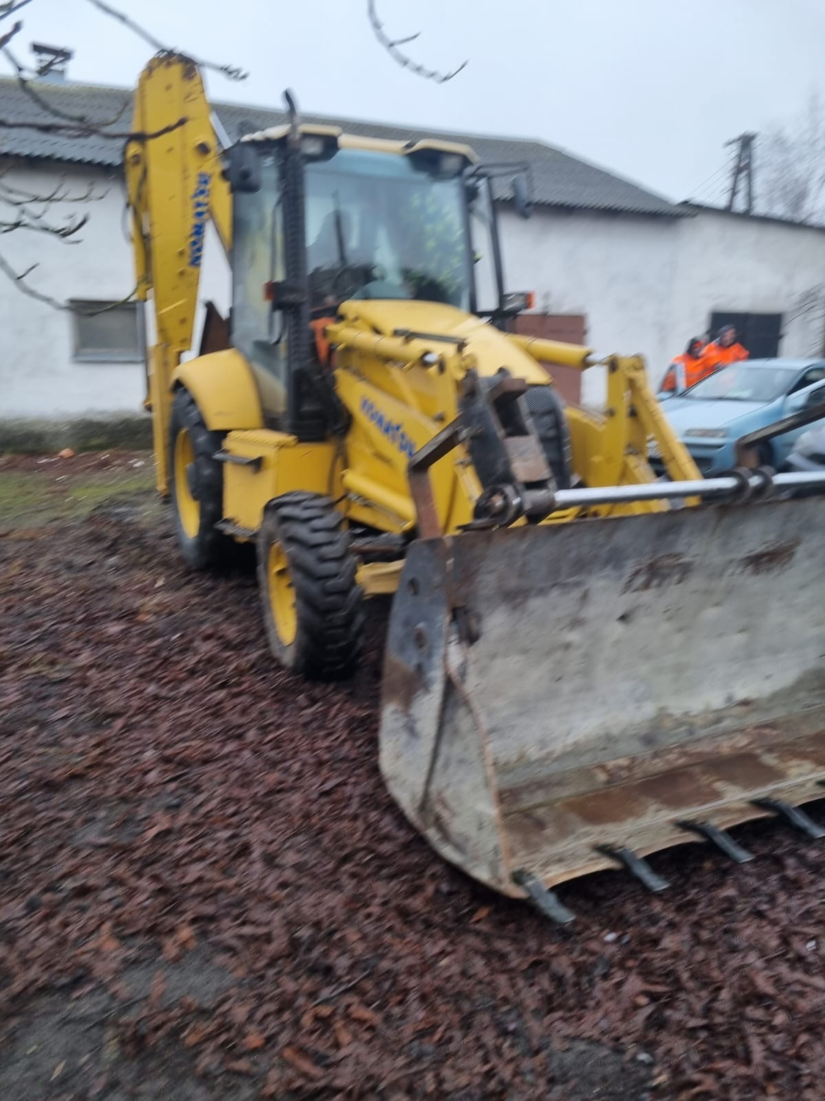 Żółta koparko-ładowarka Komatsu na zewnątrz, z widocznym łyżką i tylnym ramieniem, na tle białego budynku i dwóch osób w pomarańczowych kurtkach.