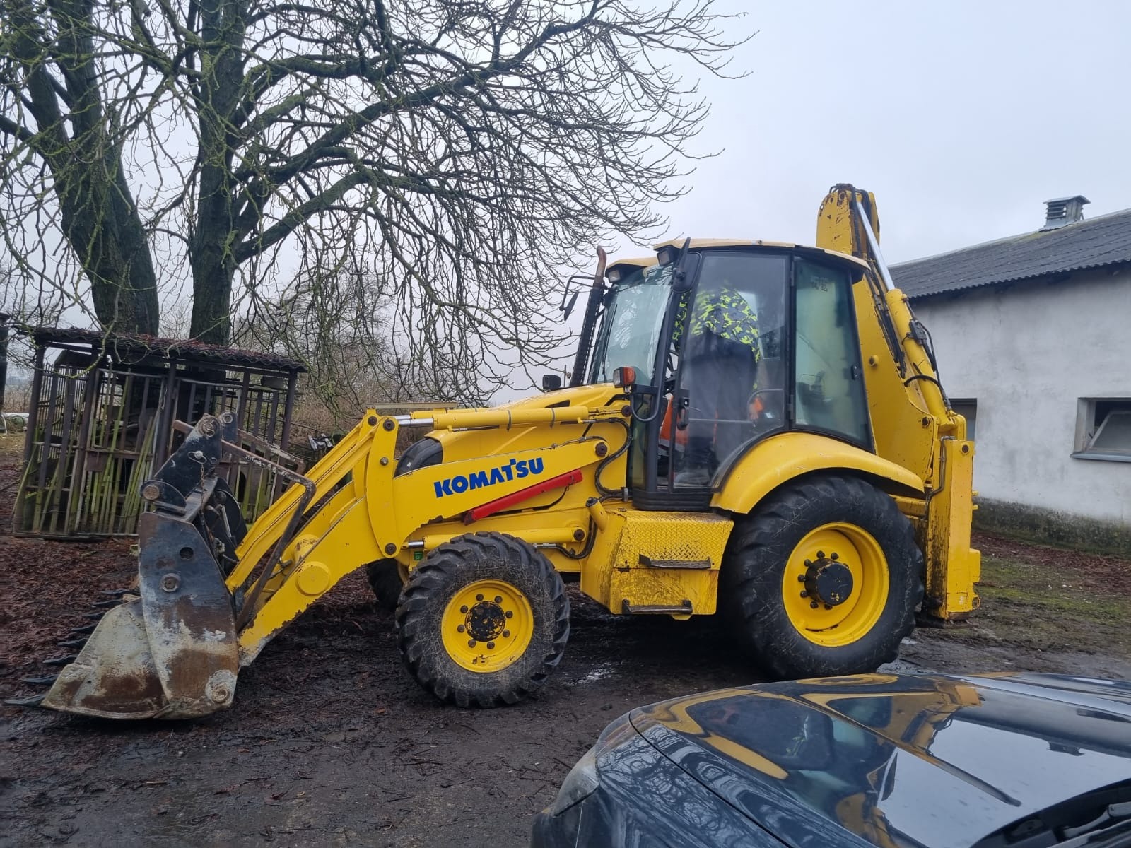 Żółta koparko-ładowarka Komatsu na błotnistym terenie, z widocznym operatorem w kabinie, na tle drzewa bez liści i budynku gospodarczego.