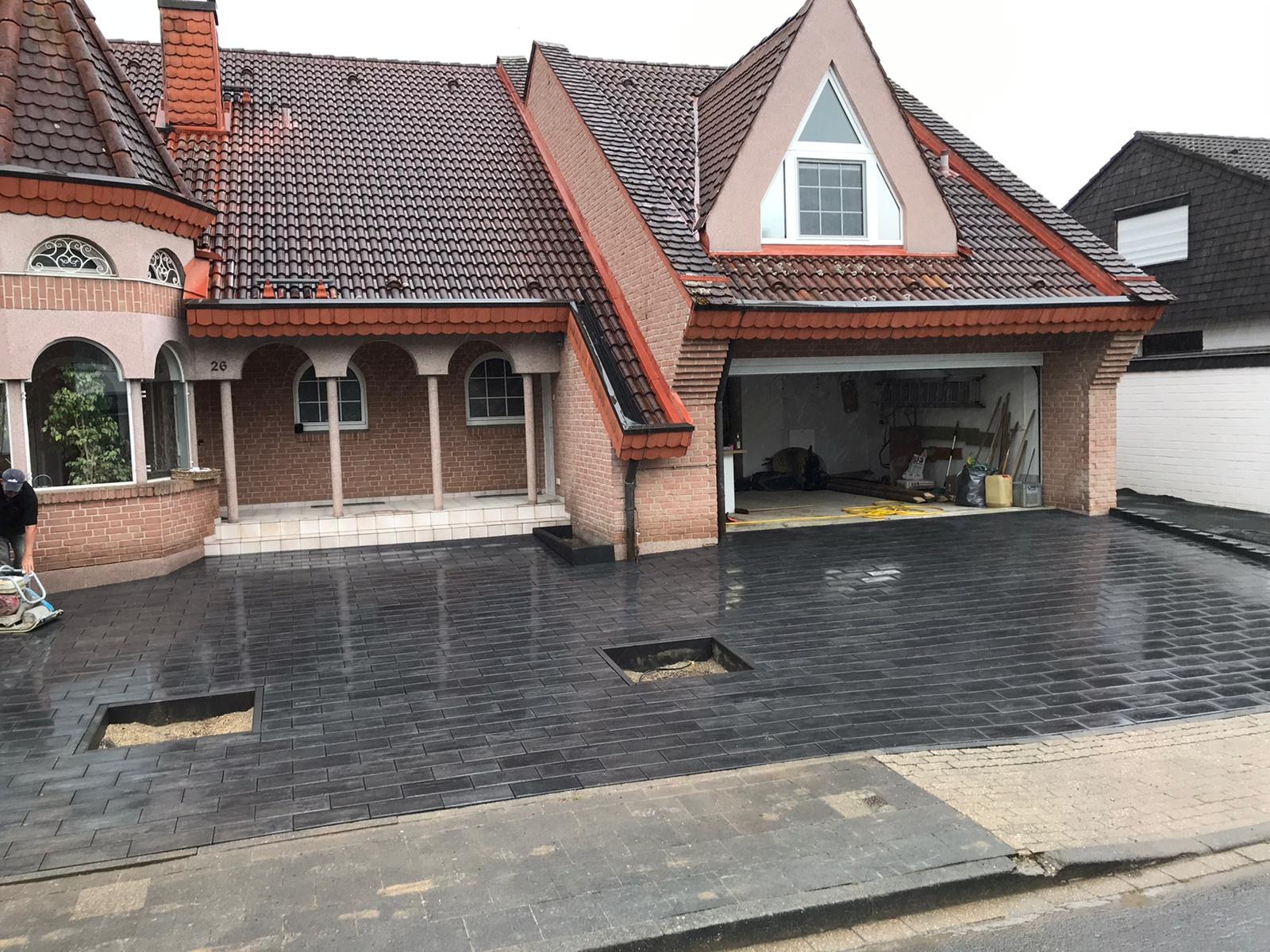 Ułożona, mokra, grafitowa kostka brukowa na podjeździe przed domem z odsłoniętymi fragmentami podłoża w dwóch miejscach, widoczny pracownik z maszyną wibracyjną, brama garażowa otwarta.