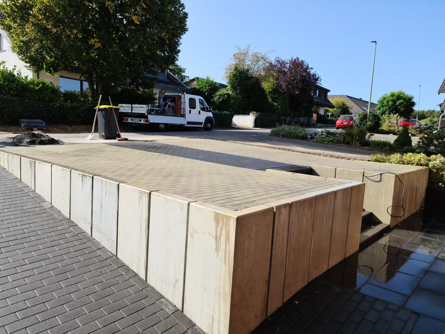 Ułożona kostka brukowa na podjeździe, widoczne betonowe obrzeża, zaparkowany biały samochód dostawczy w tle, grabie i zmiotka oparta o kosz na śmieci.