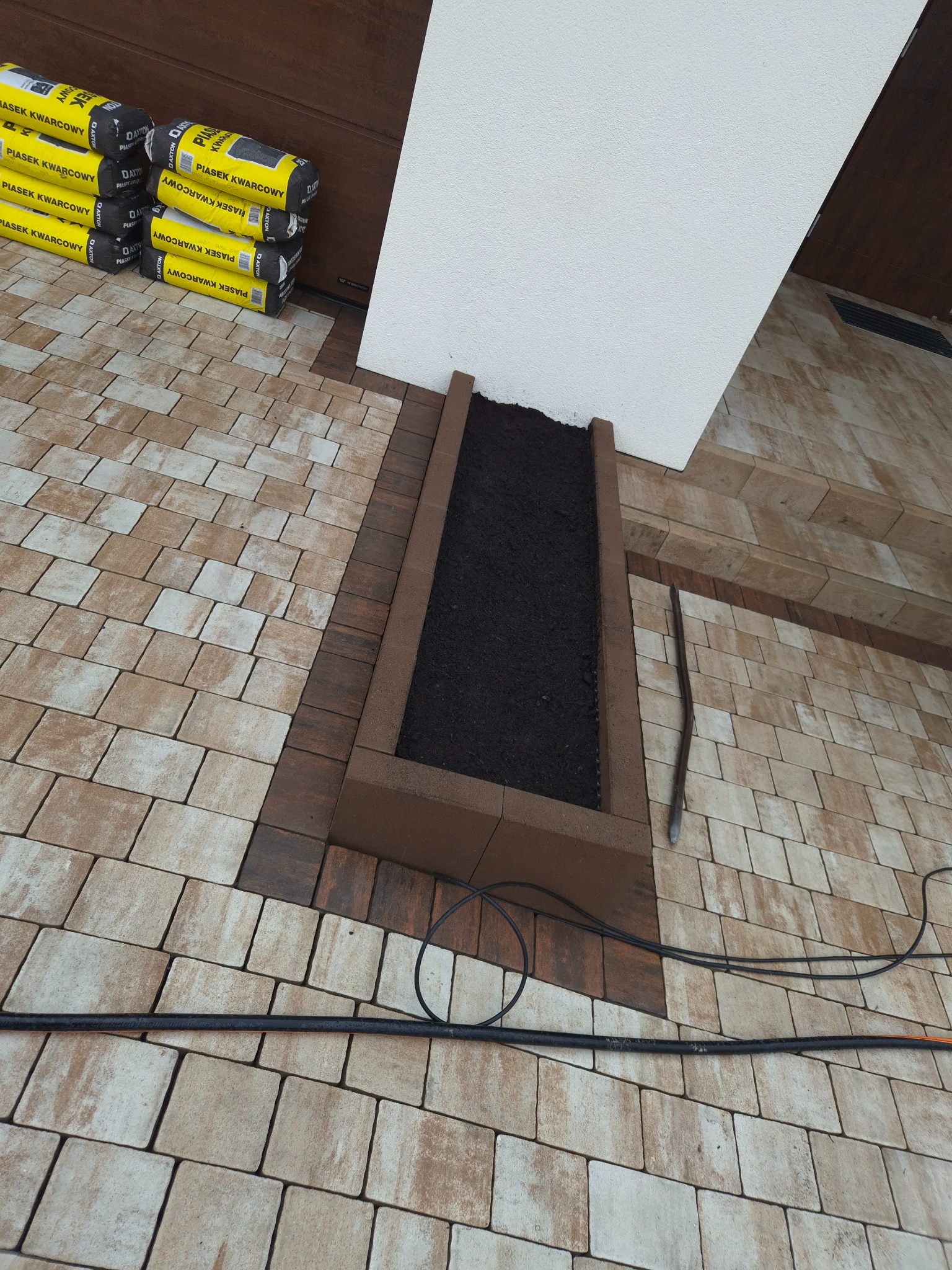 Ułożona kostka brukowa w odcieniach beżu i brązu, z wydzielonym miejscem na rabatę wypełnioną ziemią, obok zapas worków z piaskiem kwarcowym i narzędzie brukarskie
