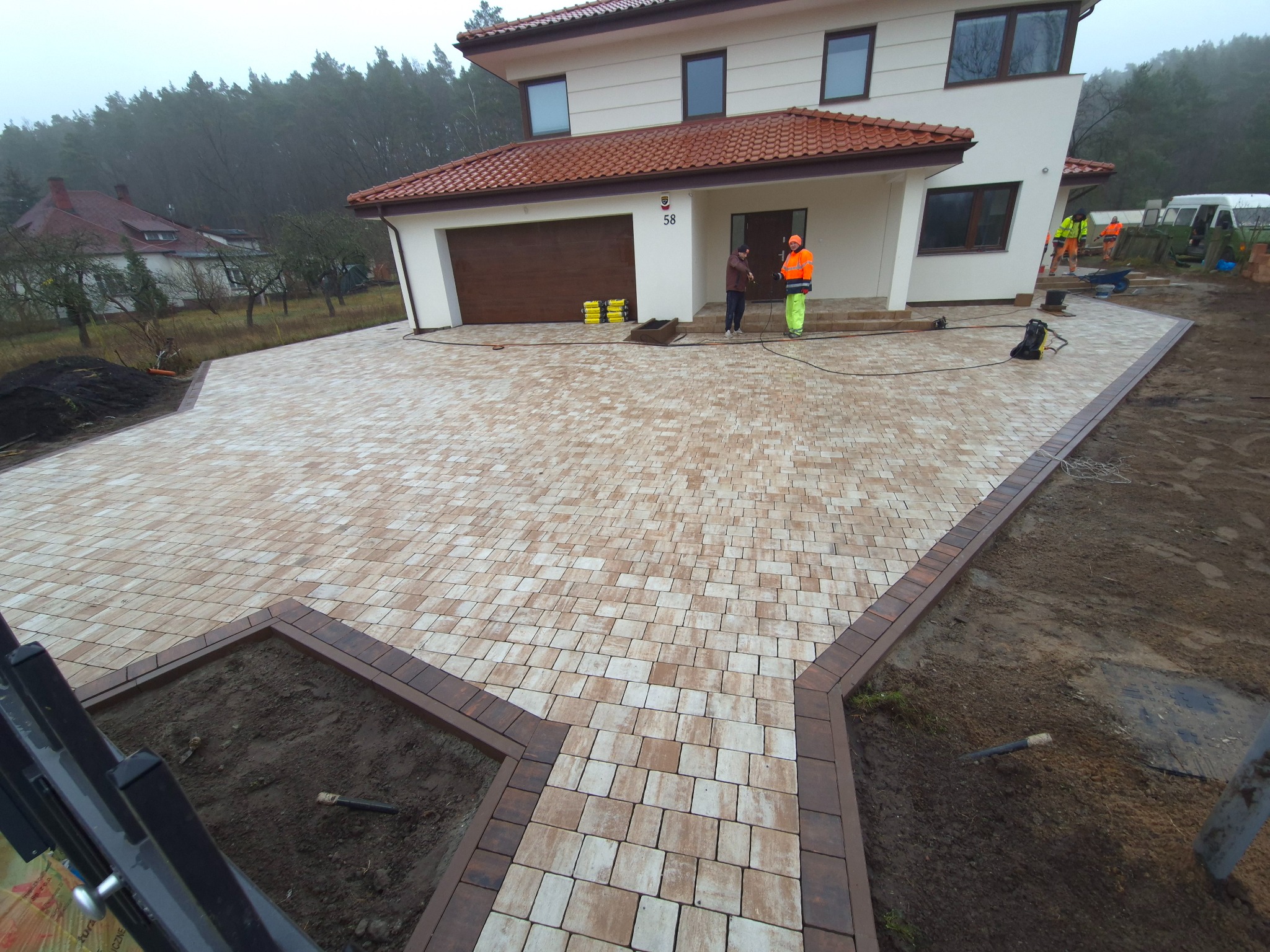 Nowo ułożona kostka brukowa w odcieniach beżu i brązu przed nowoczesnym domem z brązową bramą garażową, dwóch pracowników w odblaskowych ubraniach podczas czyszczenia kostki, widoczne elementy...