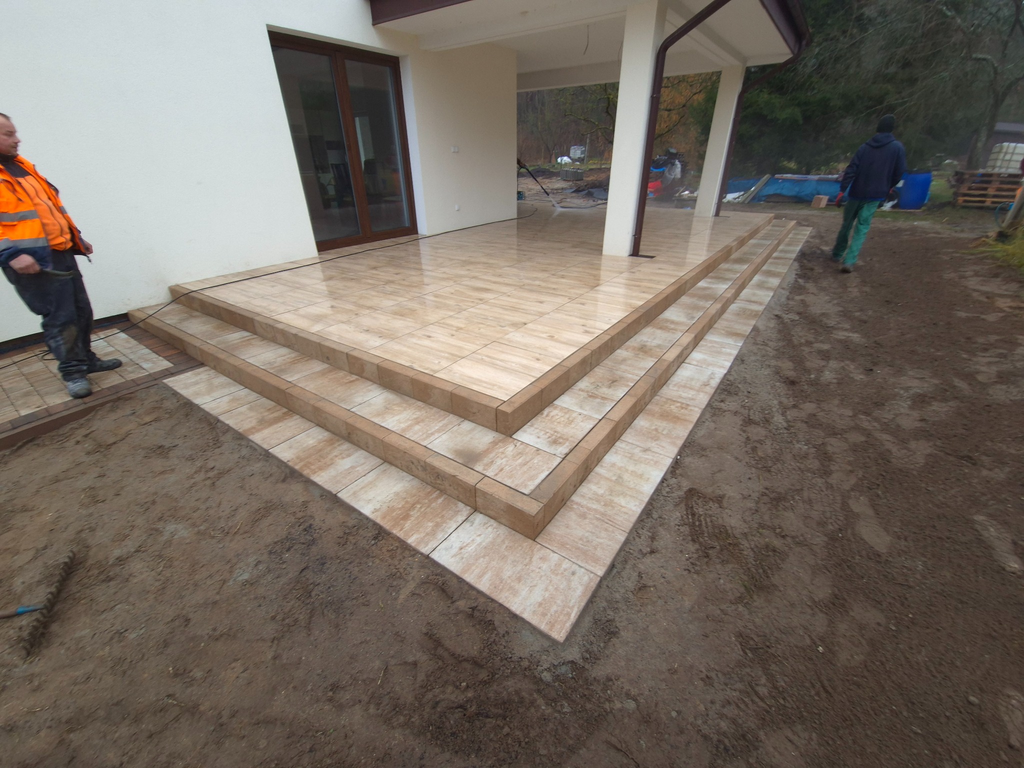 Wyłożony płytkami taras z trzema schodami przed domem, widoczny pracownik w odblaskowej kamizelce i przygotowany teren wokół.