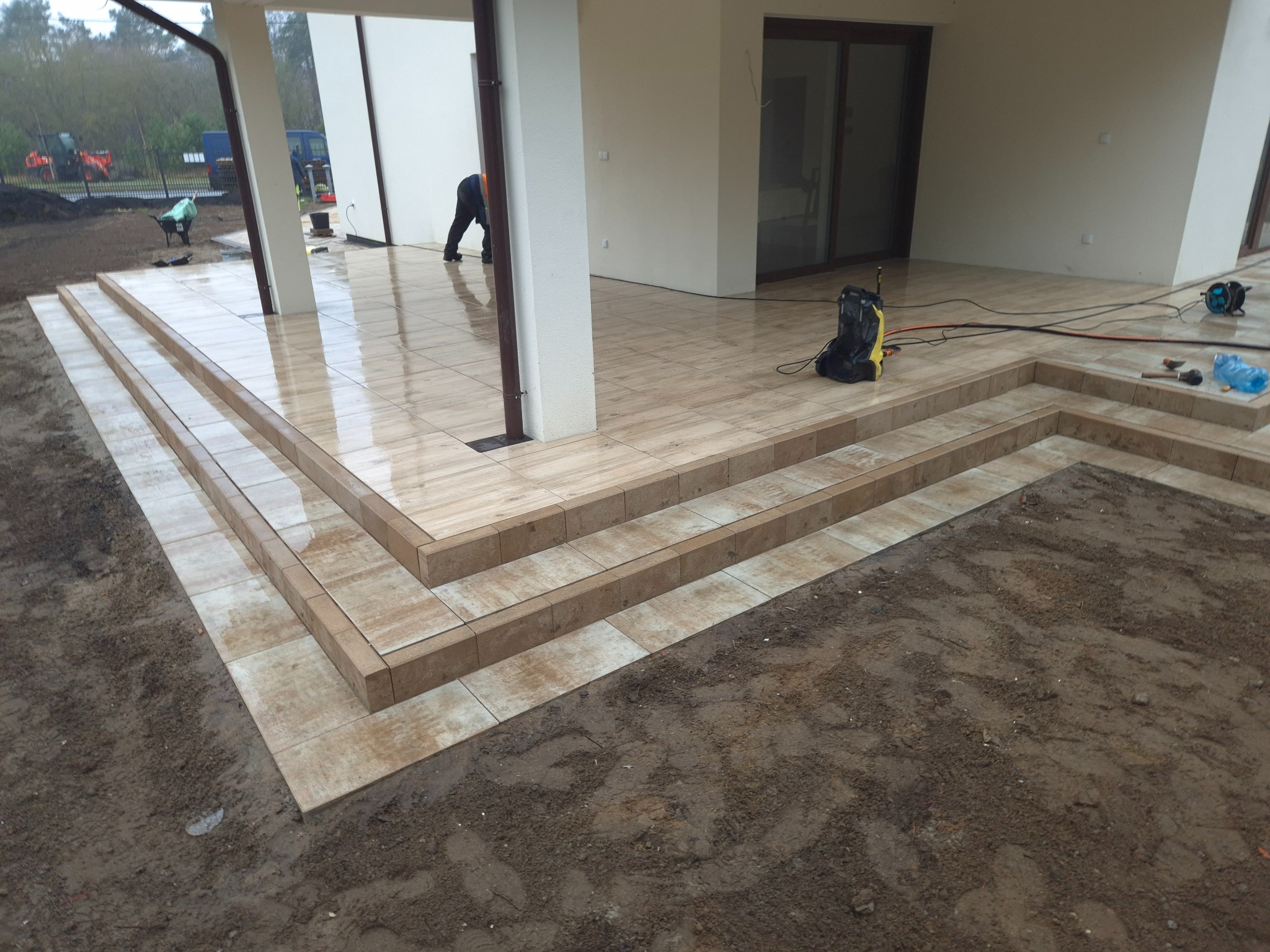 Wyłożone płytkami schody tarasowe w Bydgoszczy, z widocznym fragmentem tarasu i pracującym fachowcem w tle, otoczone mokrą ziemią.