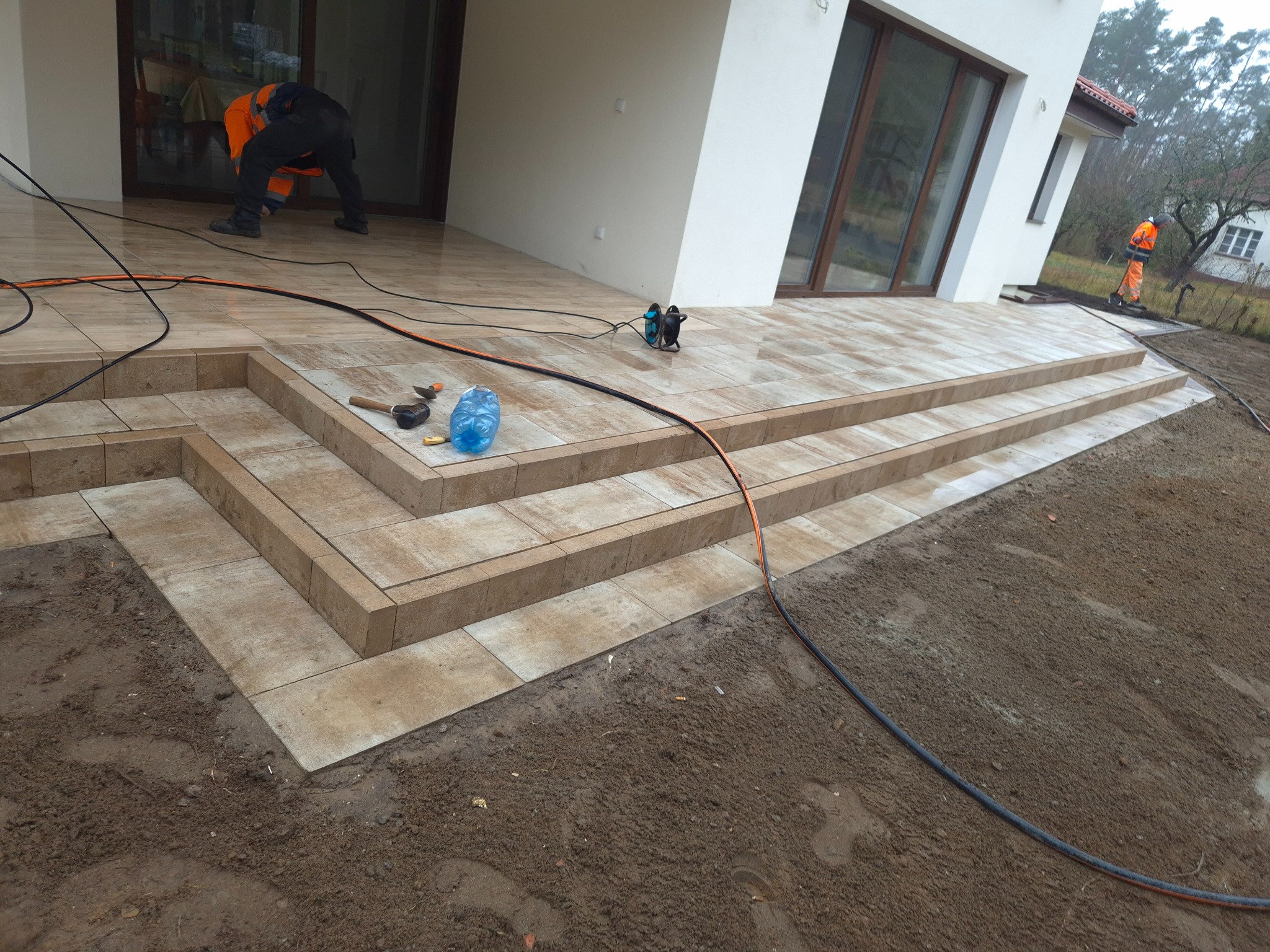 Ułożona z jasnych płytek strefa tarasowa z trzema schodami, pracownik w pomarańczowym kombinezonie wykańcza krawędzie, widoczne narzędzia i zwinięty przedłużacz.