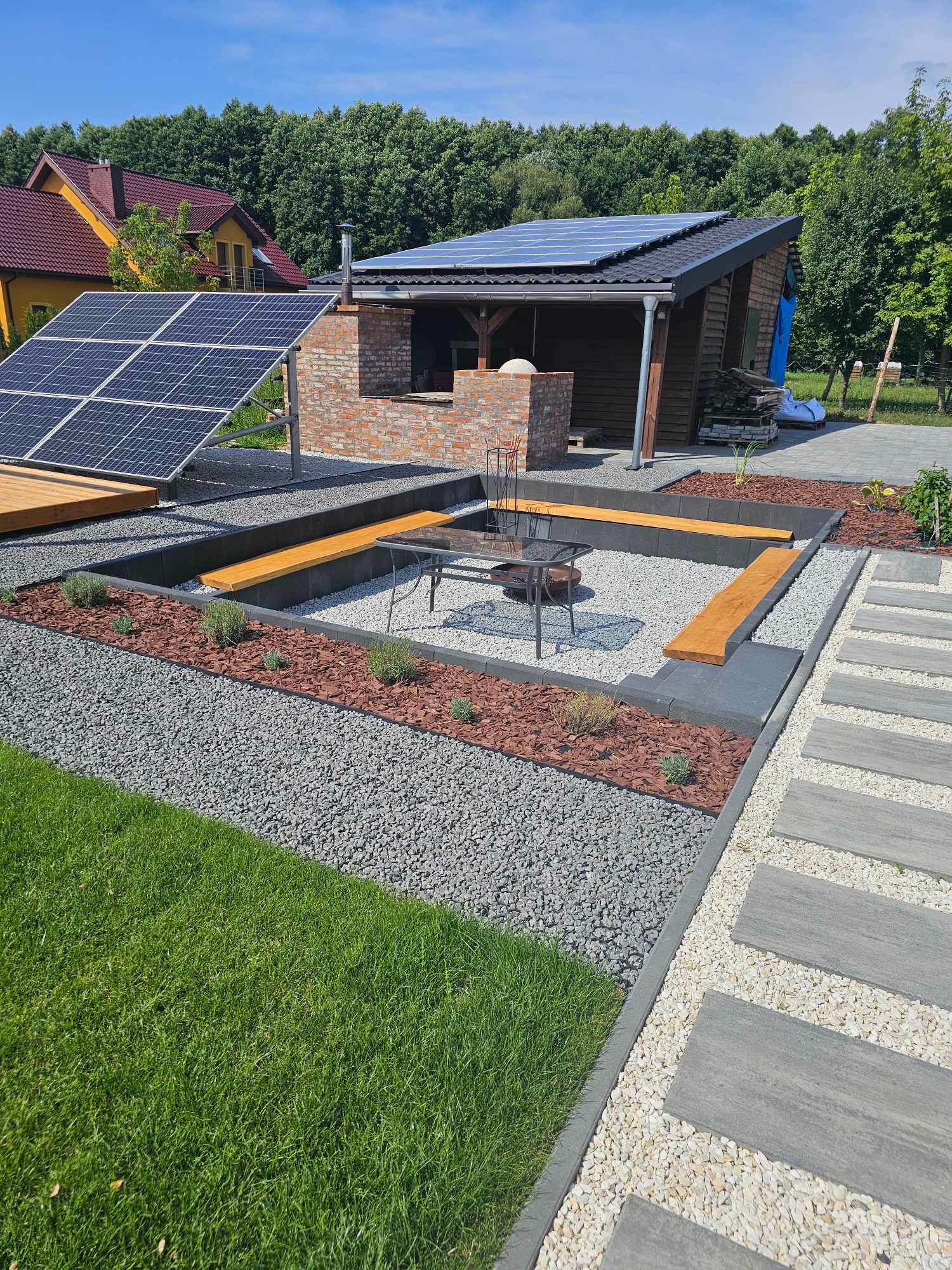 Nowoczesna aranżacja ogrodu z paleniskiem otoczonym kamieniami, drewnianymi ławkami i kostką brukową, uzupełniona o budynek z ceglanym kominkiem i panele słoneczne.